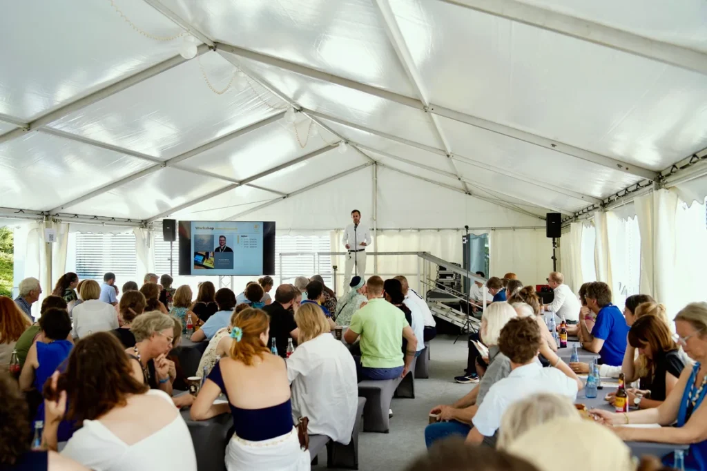 Große Besuchergruppe sitzt beim Burger Branchentreff unter einem Festzelt und verfolgt einen Fachvortrag. Der Burger Branchentreff ist ein jährliches Branchen-Event für Goldschmiede, Juweliere und Edelmetall-Profis.