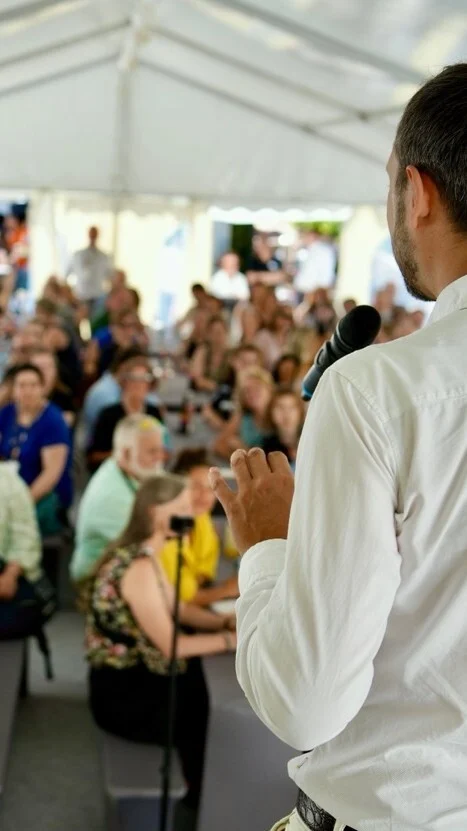 Redner mit Mikrofon auf der Bühne beim Burger Branchentreff, vor ihm sitzt ein großes Publikum aus der Edelmetallbranche. Der Branchentreff findet jährlich bei Burger Edelmetalle in Keltern statt.