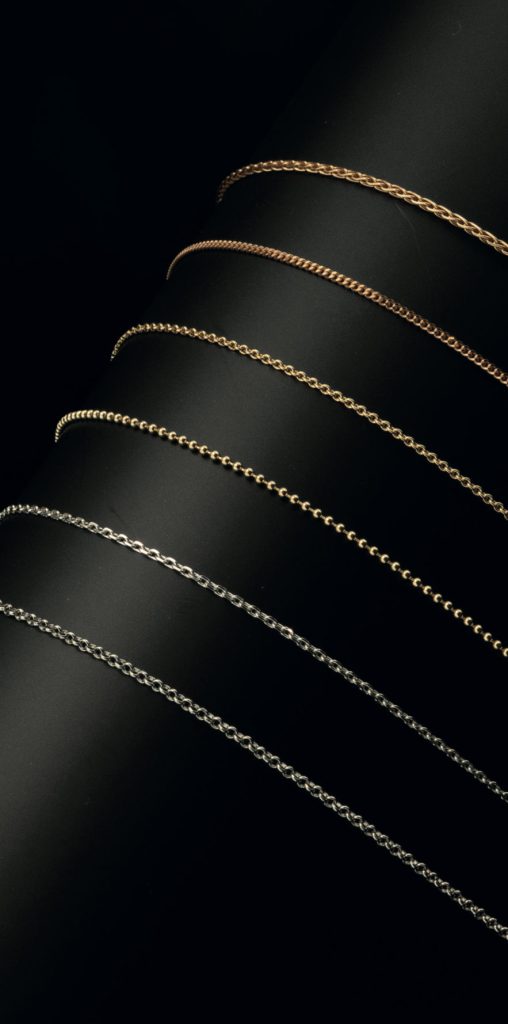 Mehrere Gold- und Silberketten in verschiedenen Kettenstilen sind spiralförmig um einen schwarzen Zylinder gewickelt und werden von unten dramatisch beleuchtet. Burger Edelmetalle bietet ein breites Ketten-Sortiment in verschiedenen Edelmetalllegierungen und Kettenstilen als Meterware und Collier.