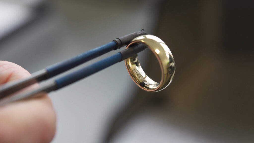 Hand hält mit Pinzette einen glänzenden Goldring bei der Gravur-Veredelung bei Burger Edelmetalle. Die hauseigene Ringfertigung bietet Gravuren, Finish und Nut in verschiedenen Edelmetallen.