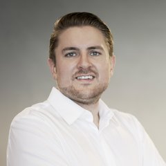 Ein Mann mit kurzem, hellbraunem Haar und gestutztem Bart, der ein weißes Button-up-Hemd trägt, steht lächelnd vor einem schlichten grauen Hintergrund in einer Scheidgutanlage.