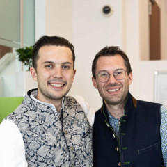 Zwei lächelnde Männer mit Edelmetall-Westen und Brillen stehen dicht beieinander und posieren für ein Foto in einem hell erleuchteten modernen Raum.