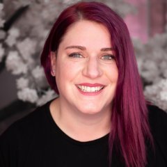 Eine Frau mit glattem, dunkelrosafarbenem Haar und schwarzem Hemd lächelt vor einem unscharfen Hintergrund mit weißen Blumen, der an die Reinheit von Scheidgut und Edelmetalle erinnert.