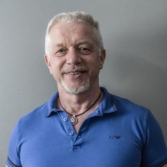 Ein lächelnder älterer Mann mit kurzen grauen Haaren und einem Bart, der ein blaues Polohemd und eine Halskette trägt, steht vor einer schlichten grauen Wand in der Scheideanstalt.