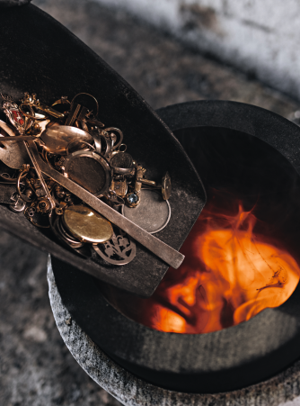 Goldschmuck, Uhren und weitere Edelmetallgegenstände werden in einer Schaufel in einen Schmelztiegel mit orangefarbenen Flammen geschüttet. Burger Edelmetalle nimmt Scheidgut aller Art entgegen und schmelzt es in der hauseigenen Scheideanstalt ein, um die Edelmetalle zurückzugewinnen.