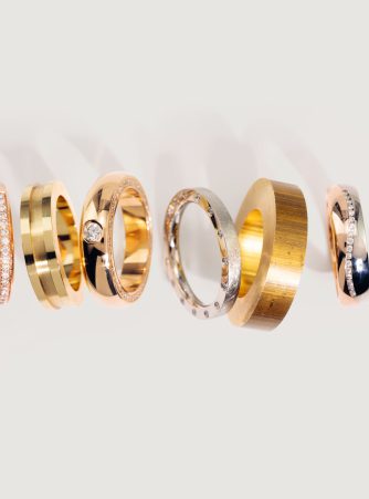 Sechs finierte Edelmetall-Ringe von Burger in Gold, Roségold und Silber, darunter Modelle mit Steinbesatz, in einer Reihe auf hellem Hintergrund. Vom Rohling bis zum finierten Trauring fertigt Burger Edelmetalle nach individuellen Vorgaben.