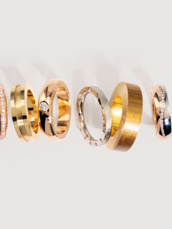 Sechs finierte Edelmetall-Ringe von Burger in Gold, Roségold und Silber, darunter Modelle mit Steinbesatz, in einer Reihe auf hellem Hintergrund. Vom Rohling bis zum finierten Trauring fertigt Burger Edelmetalle nach individuellen Vorgaben.
