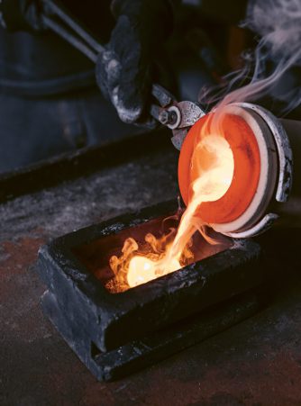 Mitarbeiter von Burger Edelmetalle gießt in Schutzhandschuhen glühendes Edelmetall aus einem Schmelztiegel in eine Barrenform, begleitet von Dampf und Flammen. Das zurückgewonnene Scheidgut wird als Edelmetallbarren oder Granulat vergütet.