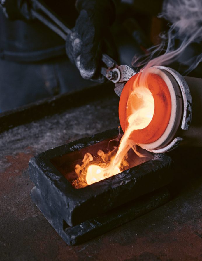 Mitarbeiter von Burger Edelmetalle gießt in Schutzhandschuhen glühendes Edelmetall aus einem Schmelztiegel in eine Barrenform, begleitet von Dampf und Flammen. Das zurückgewonnene Scheidgut wird als Edelmetallbarren oder Granulat vergütet.