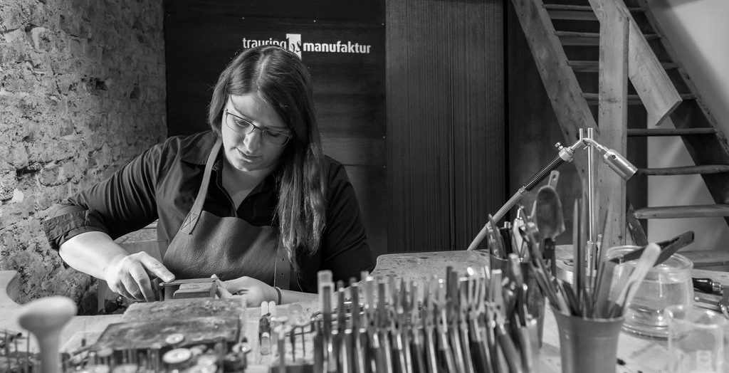 Eine Frau mit Brille und Schürze arbeitet konzentriert an einem Handwerkertisch, umgeben von Werkzeugen, und veredelt Edelmetall in einer rustikalen Werkstatt.