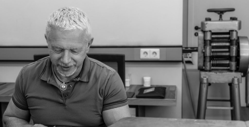 Ein Mann mit kurzen grauen Haaren arbeitet an einem Schreibtisch in einer Werkstatt der Scheideanstalt, wobei im schwarz-weißen Hintergrund Werkzeuge und Maschinen zu sehen sind.