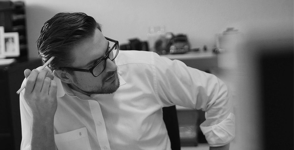 Ein Mann mit Brille und weißem Hemd sitzt an einem Schreibtisch in einem Büro der Scheideanstalt, hält sich einen Stift an den Kopf und wirkt in dieser Schwarz-Weiß-Kulisse nachdenklich.